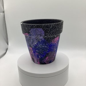 May include: A black terracotta planter with a speckled finish, painted with pink, blue, and purple abstract designs.
