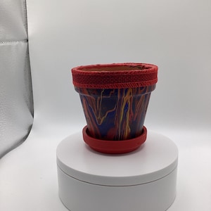 Puede incluir: Un pequeño macetero de terracota con un diseño marmoleado rojo y azul. El macetero tiene un borde rojo y una base roja.