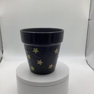 May include: A black painted flower pot with a textured surface. The pot is decorated with various sized gold and green stars. The pot is sitting on a white, round platform.