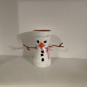 Könnte beinhalten: Ein weiß bemalter Terrakotta-Topf-Schneemann mit einem rot-weiß gestreiften Schal, einer orangefarbenen Karottennase und schwarzen Augen und Knöpfen. Der Schneemann hat braune Pfeifenputzerarme.
