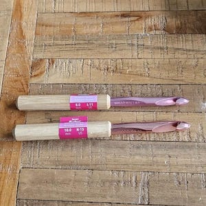 Puede incluir: Dos ganchillos con mangos de madera y ganchos rosa metálico. Uno está etiquetado 8,0 mm, L-11 US, y el otro 10,0 mm, N-15 US. Los ganchillos están sobre una superficie de madera.