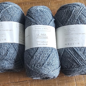 May include: Three balls of grey Le Petit Lambswool yarn, each with a white label. The labels display text including "100% Lambswool" and "Made in the UK." The yarn is arranged on a wooden surface.