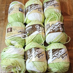 May include: Nine balls of green and white yarn are displayed. The yarn is labelled "Sugar'n Cream Stripes" and "Stripes". The yarn is self-striping, ideal for knitting and crochet projects. The yarn is arranged on a wooden surface.