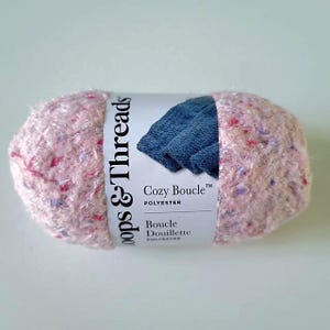 Peut inclure: Pelote de fil rose Cozy Boucle en polyester avec des mouchetures violettes et rouges. L'emballage porte une étiquette blanche avec le texte "Loops & Threads" et "Cozy Boucle". L'étiquette présente également une image bleue.