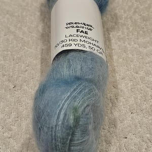 以下が含まれることがあります： 青とターコイズ色のレース糸の玉。ラベルには「December Turquoise Fae」と書かれており、糸は70/30 Kid Mohair、459 yds、50 GRと記載されています。