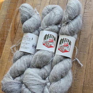 Puede incluir: Tres madejas de hilo gris claro con etiquetas. Las etiquetas presentan un diseño rojo y blanco con el texto "Dimond Laine" y una ilustración de un bosque. El hilo es suave y esponjoso, perfecto para proyectos de tejido o ganchillo.