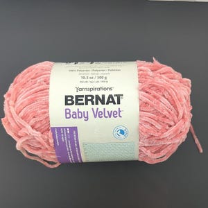 Puede incluir: Ovillo de lana rosa Bernat Baby Velvet. La etiqueta dice "10.5 oz / 300 g" y "432 yds / 450 m". La lana es suave y afelpada, ideal para mantas de bebé y otros proyectos.
