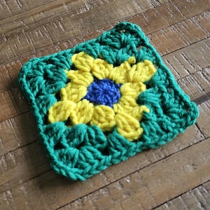 Puede incluir: Una pieza cuadrada de ganchillo hecha a mano con un diseño de girasol. El centro de la flor es azul, rodeado de pétalos amarillos. El borde es verde vibrante. Probablemente un posavasos o elemento decorativo, de unos 15 cm x 15 cm.