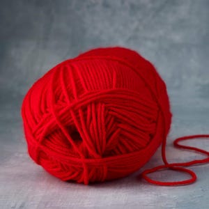 May include: A vibrant red ball of yarn, perfect for knitting or crocheting projects. The yarn is a solid, saturated color, with a soft, textured appearance. A strand of the yarn extends from the ball, suggesting its use in crafting.
