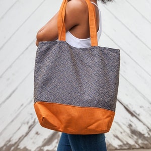 Tote bag main fabric of antique gold flourishes designer upholstery fabric on navy blue background. Straps and bag bottom of rust orange designer upholstery fabric. Model carries bag over shoulder. Blue and orange shopping bag, tote or carryall.