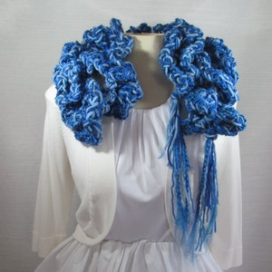 Hand crochet chunky ruffled mixed blues scarf made with multiple strands of varied texture yarns in shades of royal, sapphire and ocean blue. Scarf doubled and draped around neck with long tassels hanging down side. Ruffles surround the neckline.