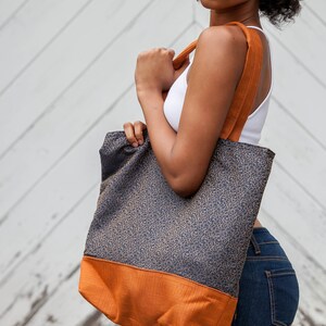 Tote bag main fabric of antique gold flourishes designer upholstery fabric on navy blue background. Straps and bag bottom of rust orange designer upholstery fabric. Model carries bag over shoulder. Blue and orange shopping bag, tote or carryall.