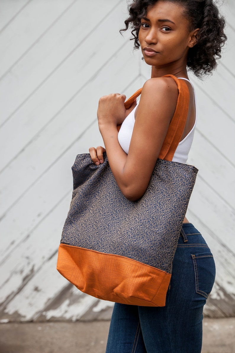 Tote bag main fabric of antique gold flourishes designer upholstery fabric on navy blue background. Straps and bag bottom of rust orange designer upholstery fabric. Model carries bag over shoulder. Blue and orange shopping bag, tote or carryall.