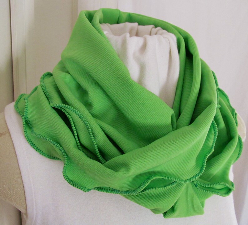 Bright green infinity scarf is folded lengthwise and wrapped double around the neck of mannequin. Lime green knit fabric scarf features kelly green machine stitched ruffled edge. Lovely spring green reminiscent of spring lettuce and greens.