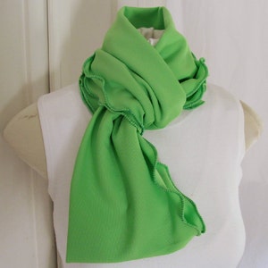 Bright green infinity circle scarf is folded lengthwise, tied around neck with one end tucked through the loop of other end. Lime green knit fabric scarf features kelly green machine stitched ruffled edge. Remiscent of spring lettuce greens.