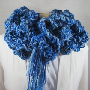 Mixed blues crochet scarf doubled and looped around neck with tassels through loop and hanging down front. Ruffles fill the neckline. Close up view shows various shades of blue and wavy, smooth and boucle textures of yarns.