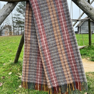 Pode incluir: Uma manta xadrez multicolorida, predominantemente cinza, vermelha e bege, com uma borda com franjas. A manta está pendurada em uma estrutura de madeira ao ar livre. O fundo apresenta um balanço e um pequeno edifício de madeira. O padrão é um xadrez clássico.