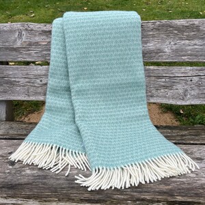 Peut inclure: Une couverture pliée, bleu clair, avec un tissage texturé et une frange blanche. La couverture est drapée sur un banc en bois vieilli, mettant en valeur sa texture douce et son design. La frange pend des bords de la couverture.