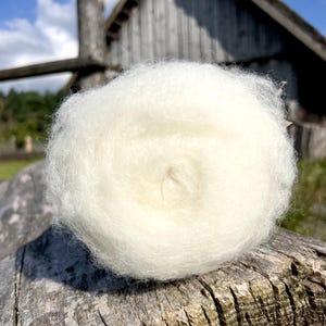 May include: A ball of soft, white wool, likely for crafting. The wool is fluffy and appears freshly processed. The background includes a weathered wooden structure and a blue sky, suggesting an outdoor setting.