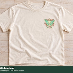 Pu&ograve; includere: Una t-shirt a maniche corte color crema con una piccola grafica sulla parte superiore sinistra del petto. La grafica raffigura una falena luna verde chiaro circondata da piccoli fiori rosa e arancioni e foglie verdi sopra le ali.