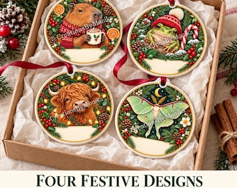 Può includere: Quattro ornamenti rotondi in ceramica in una scatola, con illustrazioni di un capibara con tazza, una rana con berretto rosso, una mucca highland e una falena verde, circondati da rami di pino, bacche rosse e nastri.