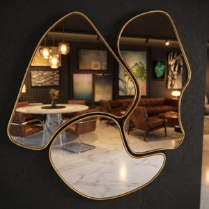 May include: A modern, abstract wall mirror with a gold-colored frame. The mirror is composed of three irregularly shaped sections, reflecting a room with furniture and artwork. The mirror is mounted on a dark gray wall, creating a striking contrast.