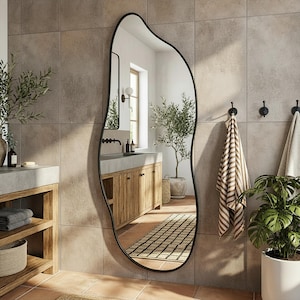 May include: An irregularly shaped, black-framed mirror hangs on a tiled wall in a bathroom. The mirror reflects a wooden vanity with a concrete countertop, a window, and a potted olive tree. Towels and a plant are also visible.