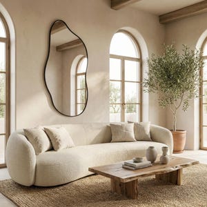 May include: A living room with a cream-coloured curved sofa and matching cushions. A large, irregularly shaped black-framed mirror hangs on the wall. A wooden coffee table and a potted olive tree complete the decor.
