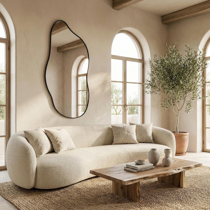May include: A living room with a cream-coloured curved sofa and matching cushions. A large, irregularly shaped black-framed mirror hangs on the wall. A wooden coffee table and a potted olive tree complete the decor.