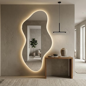 May include: An organically shaped mirror with a warm, illuminated border, mounted on a textured wall. A wooden console table with a vase and decorative items is positioned below the mirror. A black, linear pendant light hangs above the table.