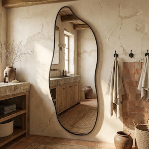 Op de afbeelding: Een grote, onregelmatig gevormde spiegel met een zwart frame domineert een rustieke badkamer. De spiegel reflecteert het interieur van de kamer, inclusief een houten wastafel, terracotta tegels en muren in neutrale tinten. Handdoeken hangen aan de muur en een geweven mand staat op de vloer.