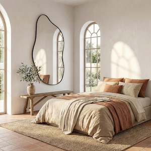 May include: A bedroom with a bed dressed in beige and terracotta bedding, including pillows and a throw. A large, black, organically shaped mirror is mounted on the wall. A wooden bench and a vase with greenery enhance the room's decor. The room has arched windows.