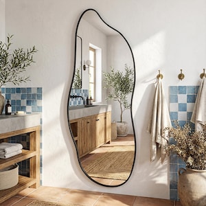 May include: An irregularly shaped, full-length mirror with a black metal frame. The mirror is in a bathroom setting with a neutral colour palette, featuring blue and white tile accents, wooden cabinetry, and a potted olive tree. The mirror's dimensions are approximately 152 cm in height.