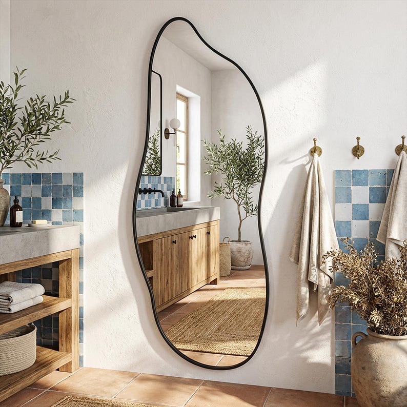 May include: An irregularly shaped, full-length mirror with a black metal frame. The mirror is in a bathroom setting with a neutral colour palette, featuring blue and white tile accents, wooden cabinetry, and a potted olive tree. The mirror's dimensions are approximately 152 cm in height.