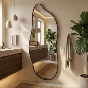May include: A large, organically shaped mirror with a black frame dominates a bathroom scene. The mirror reflects the room's features: a wooden vanity, indoor plants, and pendant lights. A neutral-toned rug is visible on the floor, adding warmth to the space.