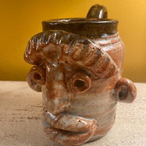 May include: A handmade ceramic mug with a face sculpted on the front. The mug is primarily tan with reddish-brown accents and a dark brown rim and handle. The face has exaggerated features, including large eyes and a prominent nose.