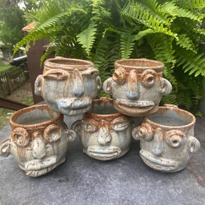 May include: A collection of six ceramic face planters. Each planter has a unique face design with brown and blue glaze. The planters are arranged on a gray surface with green foliage in the background.