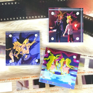 May include: Three square acrylic blocks featuring anime-style illustrations. The blocks showcase characters with spiky yellow hair and vibrant color schemes. The blocks are secured with silver-colored fasteners and are set against a backdrop of film strips.