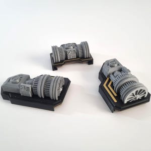 May include: Three grey miniature engine models with detailed designs, set on black bases. One is central, the others are to the sides. The models have intricate designs, suggesting a mechanical or industrial theme.