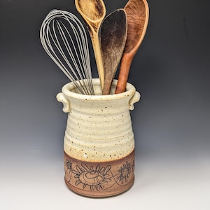 May include: A ceramic utensil crock with a speckled cream-colored top and a brown base featuring etched floral and bee designs. The vessel has two small side handles and contains a metal wire whisk and three wooden spoons of varying shades.