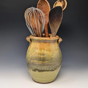 May include: A ceramic utensil crock with a mottled yellow and brown glaze, featuring two small side handles. The crock is filled with a collection of kitchen tools, including a metal wire whisk and several wooden spoons and spatulas.