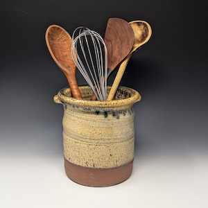 May include: A cylindrical ceramic utensil holder with a speckled beige and brown glaze, featuring two small side handles. Inside are several wooden kitchen spoons and a metal wire whisk, arranged upright within the vessel's wide opening.