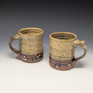 May include: Two ceramic mugs featuring a speckled tan glaze on the upper portion and a solid reddish-brown base. The lower section is decorated with a dark vine pattern and small white dots. Each mug has a curved handle and a cylindrical body.