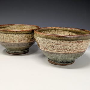 May include: Two handmade ceramic bowls. The bowls have a speckled, textured surface with bands of color. The rim is a dark brown, with a band of green and tan. The lower portion of the bowl is a darker green. The bowls are on a white surface.