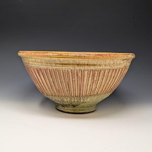 May include: A ceramic bowl featuring a wide, flared rim and a tapered base. The exterior surface displays a textured, vertical ribbed pattern in shades of tan, cream, and muted orange, with a darker, mottled green glaze covering the bottom section.