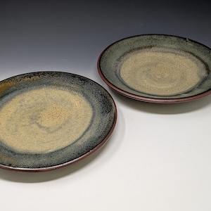 May include: Two round ceramic plates featuring a speckled beige center that transitions into a dark blue and black rim. The edges of the plates are finished with a thin, reddish-brown border, showcasing a reactive glaze with visible texture.