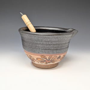 May include: A handmade ceramic mixing bowl with a spout, in shades of gray, brown, and orange. The bowl features a floral design around the base and a wooden whisk inside. This kitchenware item is perfect for mixing ingredients.