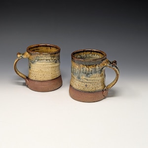 May include: Two ceramic mugs featuring a speckled cream and brown glaze on the upper portion and a solid reddish-brown base. Each mug has a curved handle and a cylindrical body with a slightly flared rim, showcasing a reactive glaze finish.