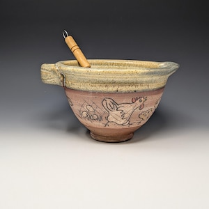 May include: A ceramic mixing bowl with a light tan rim and a pinkish-brown base featuring black line drawings of chickens and eggs. The bowl has a small handle on the left side and a pouring spout on the right, with a wooden pestle resting inside.