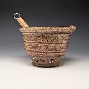 May include: A ceramic mortar with a speckled brown and tan glaze, featuring a textured exterior with horizontal ridges. The base is decorated with a hand-painted black vine pattern and small white dots. A wooden pestle with a metal loop is included.